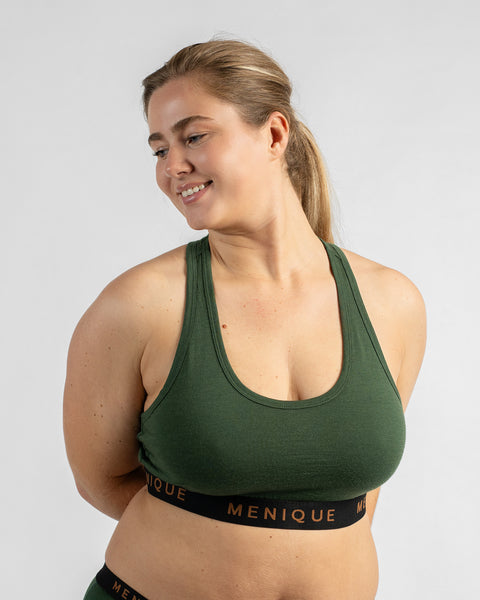 Front three-quarter view of a model looking down and smiling with her arms behind her back, wearing a forest green sports bra featuring a black elastic band with text.