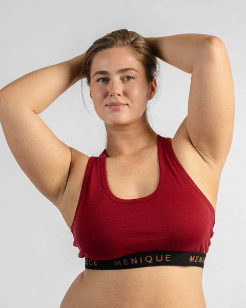 Front view of a model holding her hair up with both hands, wearing a burgundy sports bra featuring a black elastic band with text.
