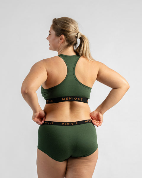 Back view of a model with her hands on her hips, wearing a forest green sports bra and matching boy-short underwear, both featuring a black elastic band.