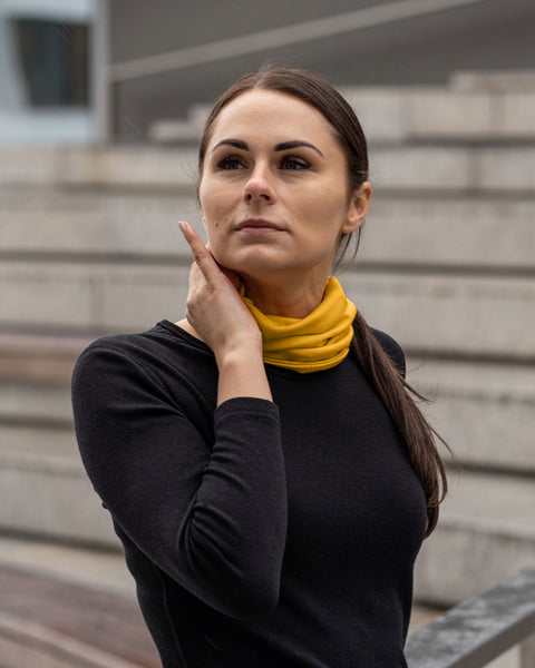A woman with dark hair pulled back in a low ponytail is looking upwards and to her left, with her right hand gently touching her neck. She is wearing a spicy yellow menique merino wool neck gaiter and a black long-sleeved shirt. The background shows a set of light-colored concrete steps and a blurred building with windows.