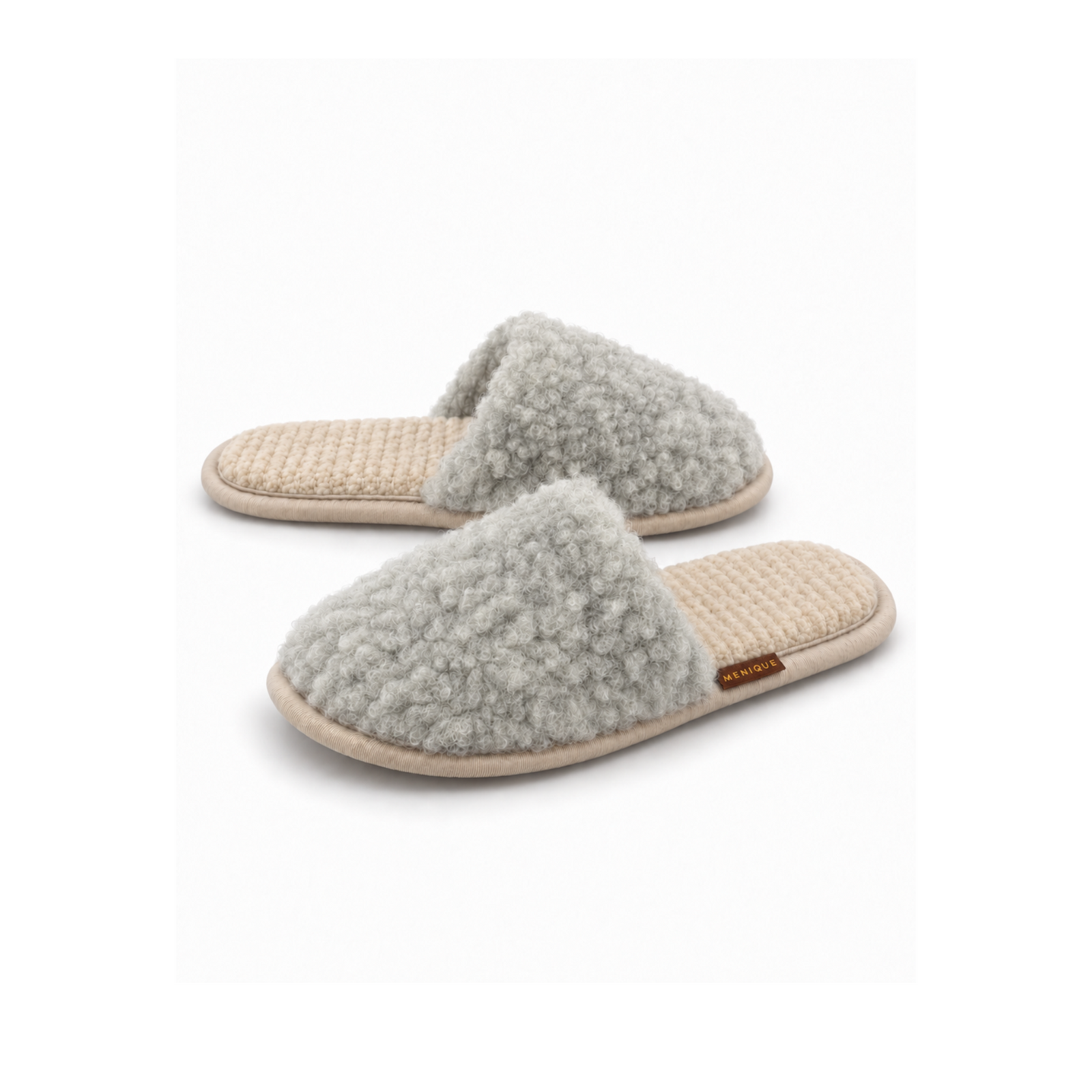A person sitting on a bed wearing beige, fuzzy slip-on slippers with a small brown "MENIQUE" tag, resting their feet on a textured rug.