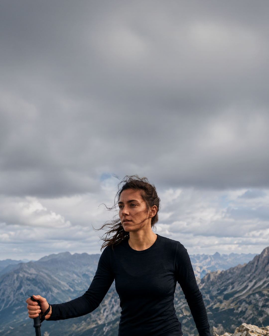 A woman in a black long-sleeve top and olive green pants hiking with a trekking pole along a rocky mountain ridge, with dramatic, jagged mountain peaks and a cloudy sky in the background.