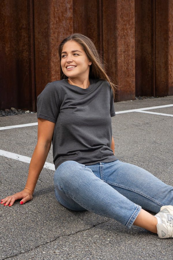 A smiling woman sitting on a paved surface, leaning back on her right arm and looking off to the side. She is wearing a charcoal grey crew neck t-shirt, blue jeans, and light grey sneakers, with a rusty corrugated metal wall in the background.