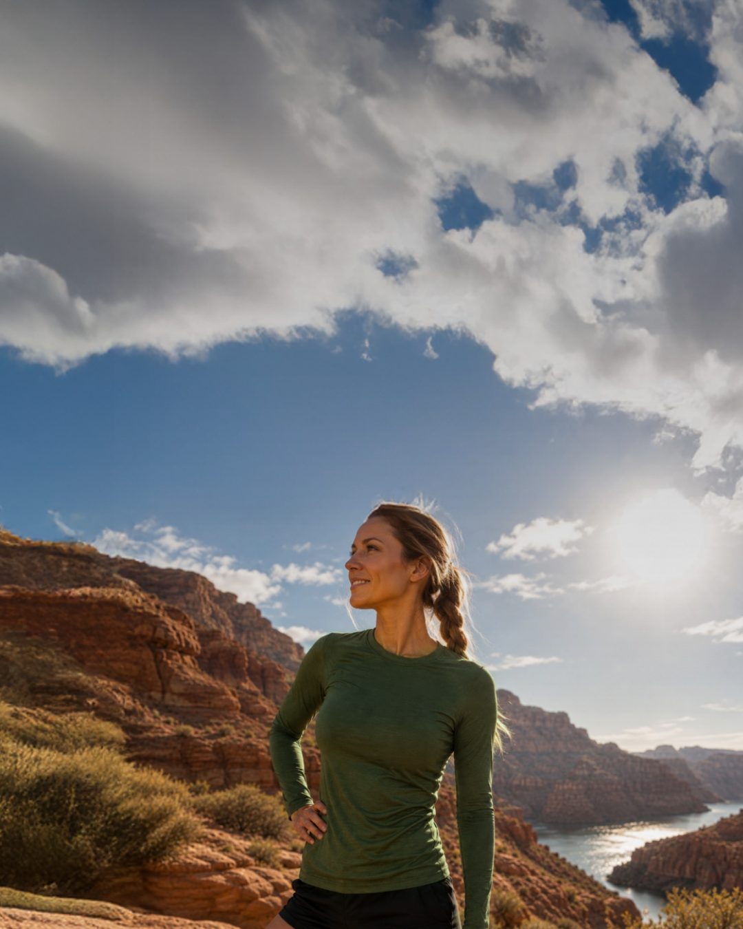 A smiling woman wearing a dark green long-sleeve shirt and black shorts standing with her hand on her hip on a rocky cliff overlooking a scenic river and canyon.
