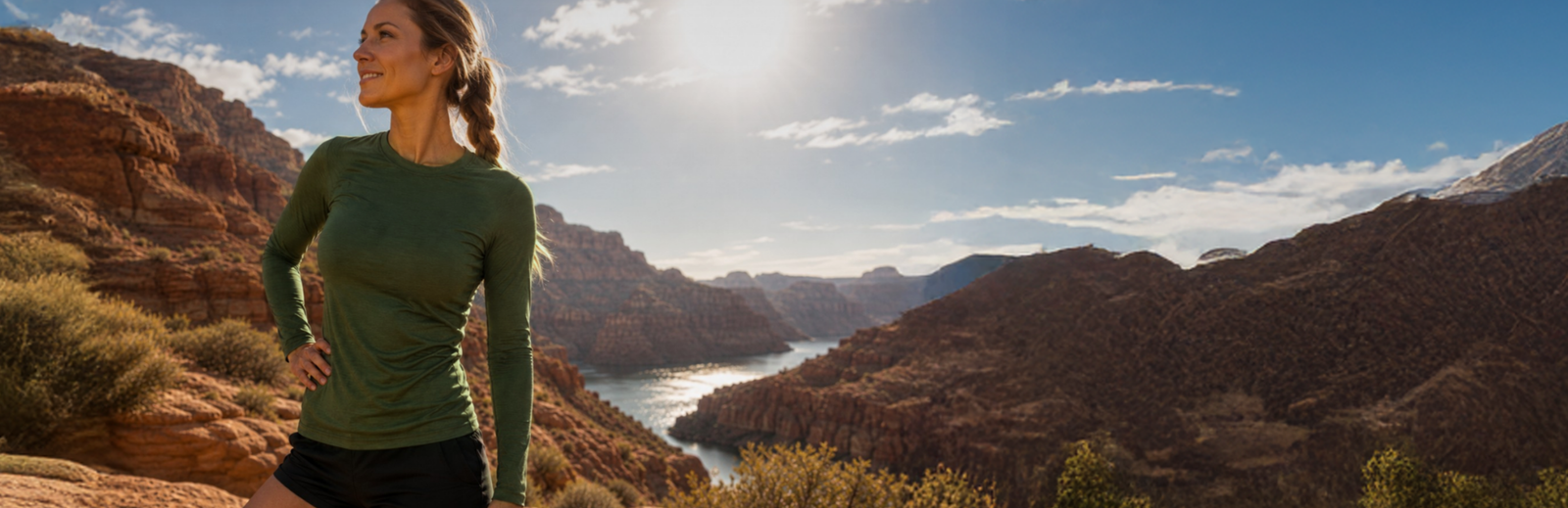 A smiling woman wearing a dark green long-sleeve shirt and black shorts standing with her hand on her hip on a rocky cliff overlooking a scenic river and canyon.