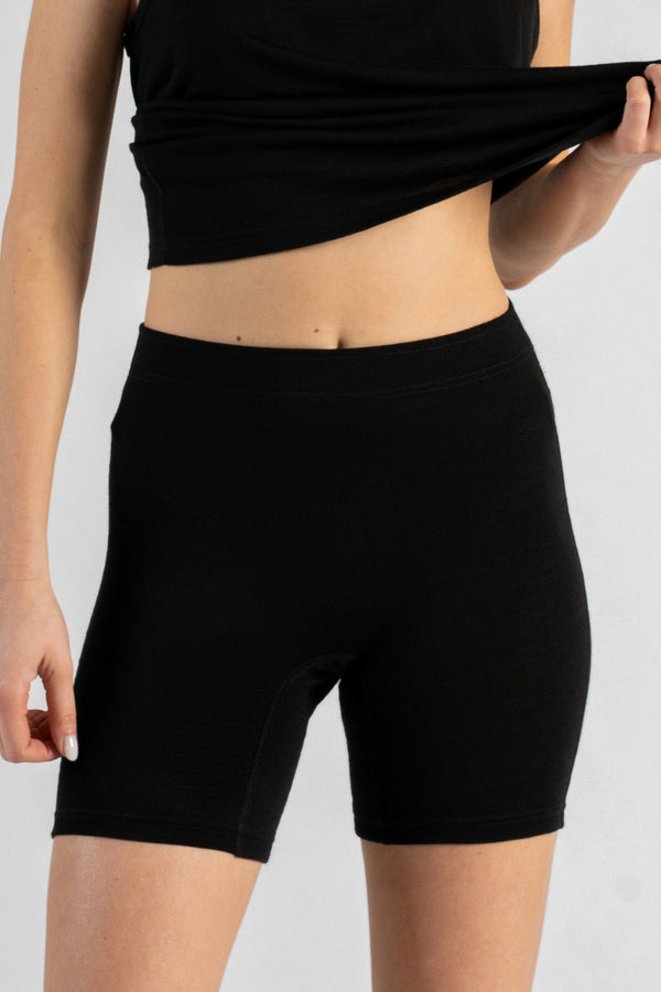 A model facing forward wearing black, form-fitting short leggings and a black top against a plain light background. Their left hand is lifting the hem of the top slightly, revealing their midriff, while their right arm hangs relaxed by their side.