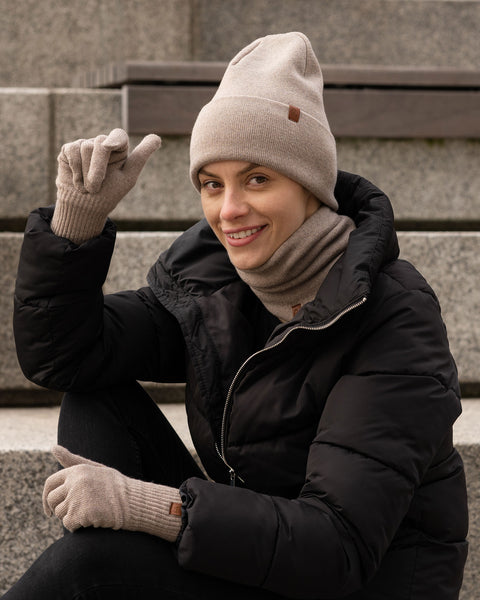 Woman wearing creamy beige Merino wool beanie, gloves, and neck gaiter while sitting outdoors on stone steps in a black winter jacket.