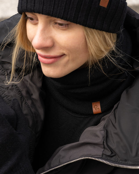 A woman wearing a black knit beanie, black knit neck gaiter, and a warm black jacket, smiling softly while outdoors in cold weather.