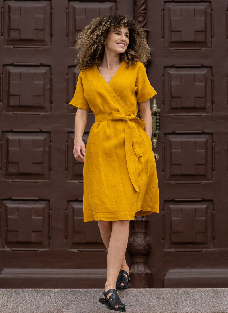 Woman wearing a mustard yellow linen wrap dress with short sleeves and a tie waist, walking confidently in front of a dark wooden architectural door. The lightweight dress highlights the natural texture of linen and offers a comfortable yet elegant silhouette for warm-weather occasions.