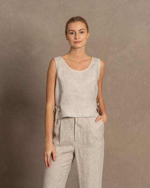 A woman with blonde hair pulled back wears a natural linen sleeveless crop top with side ties, paired with matching light natural linen pants. She stands facing forward with one hand in her pocket against a muted brown textured background.