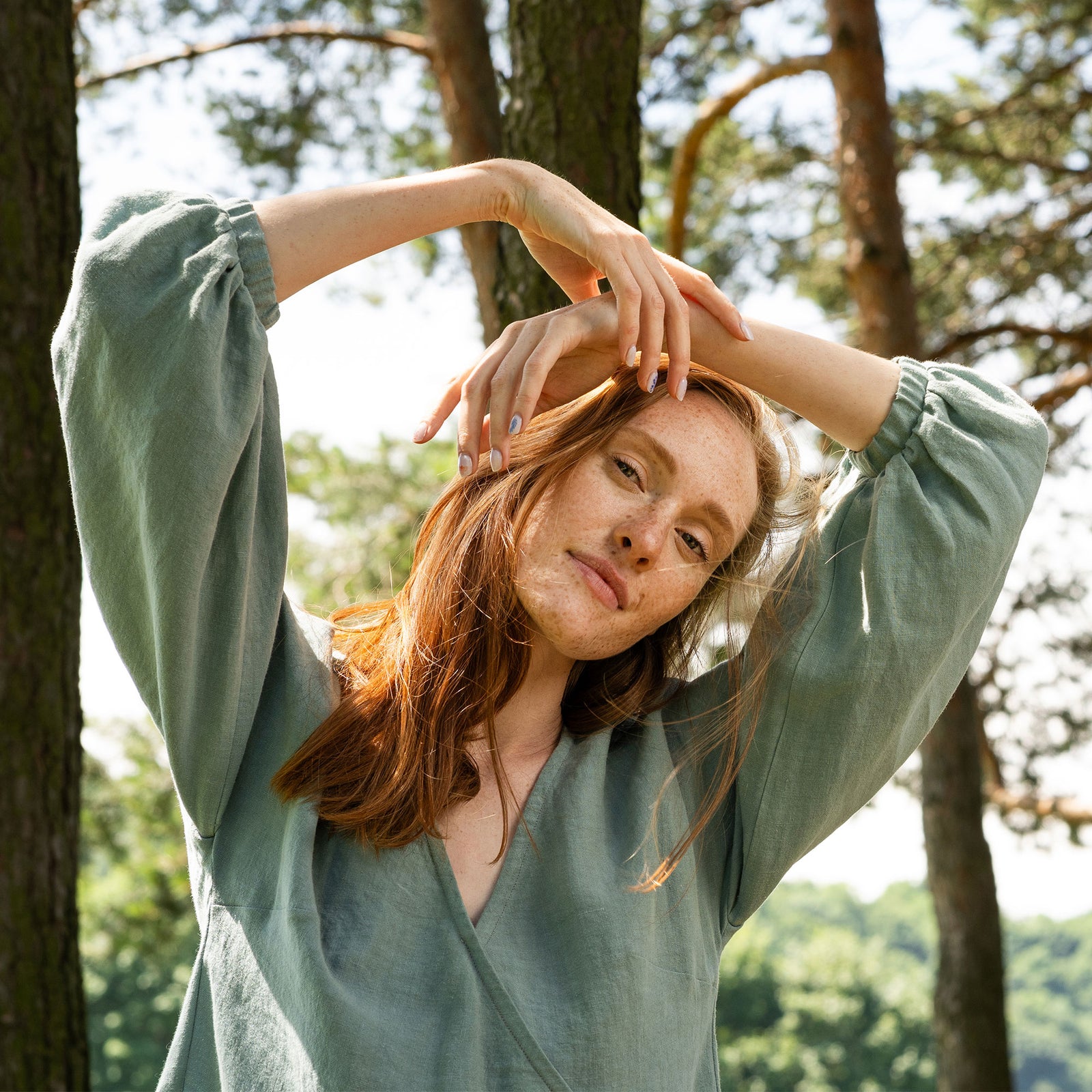 A full-body shot of a woman with long, reddish-brown hair standing in a grassy forest. She is looking upwards and to her right. She is wearing a knee-length, mint green linen wrap dress with a V-neck and elbow-length, puffed sleeves. She is holding a woven basket filled with white flowers and green foliage in her left hand. She is wearing black slides. 