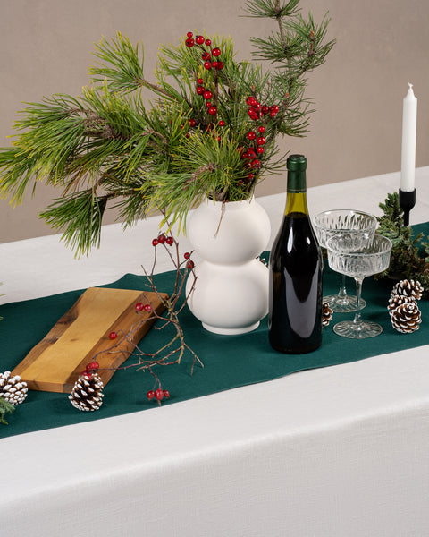 The menique linen table runner in dark green color, decorating a Christmas dinner table. There is a wine bottle, glasses, and a wooden table on top of the table top. there is also a white linen tablecloth underneath the runner.
