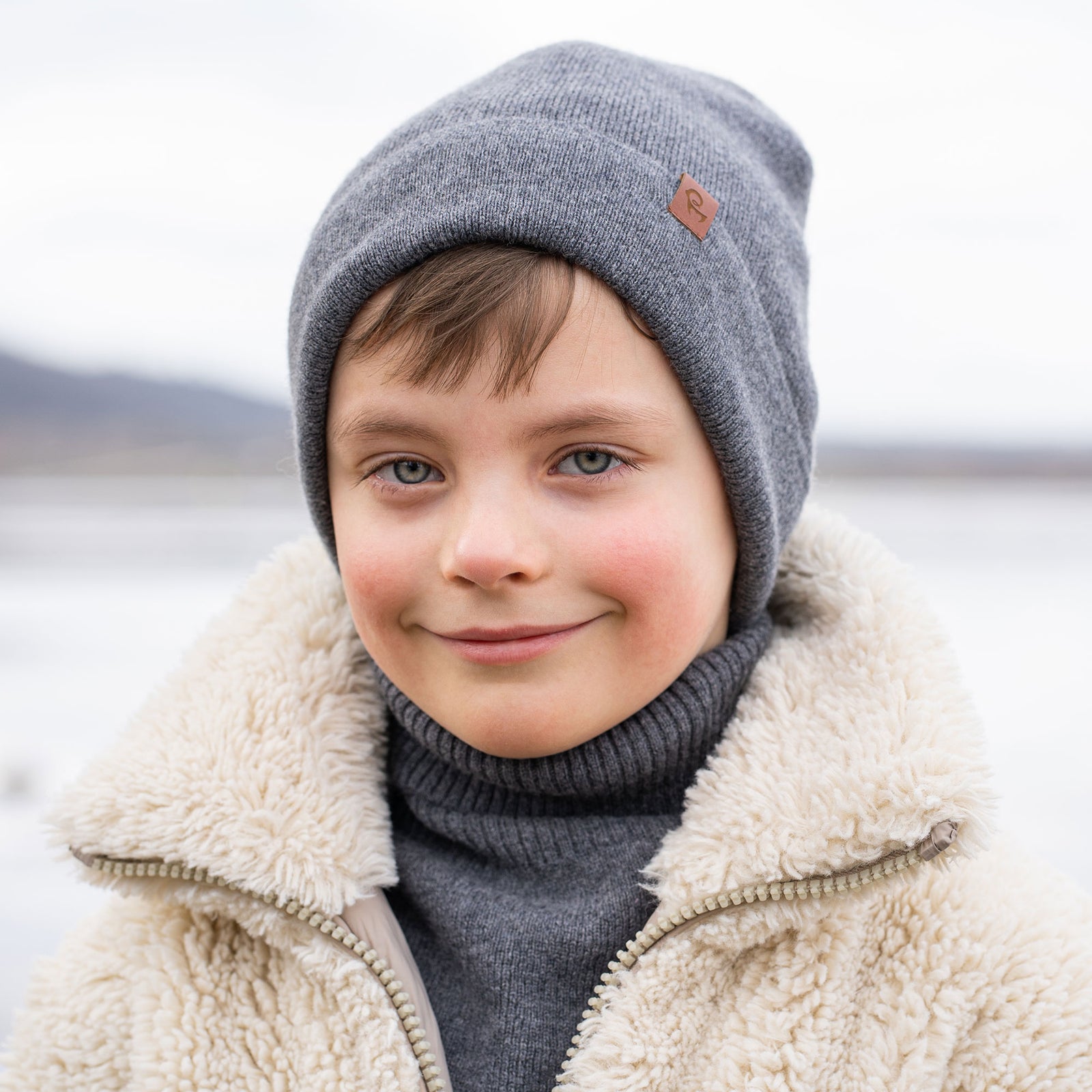 A young child with short brown hair and a wide smile, is dressed in winter clothing. They are wearing a beige beanie, a beige turtleneck sweater, and a cream-colored sherpa jacket, with beige knitted gloves. A snowy, blurred background with what appears to be a body of water is visible behind them.
