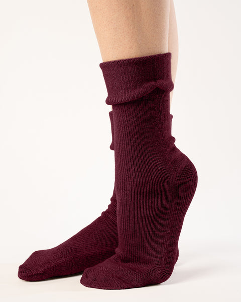 A side view captures a person from the mid-thigh down, showcasing ribbed, royal cherry socks with a partially folded cuff. The socks reach the mid-calf, and the person&#39;s feet are positioned with one slightly behind the other, heel raised. They stand against a stark white backdrop with even lighting that accentuates the texture and gathered folds of the socks.