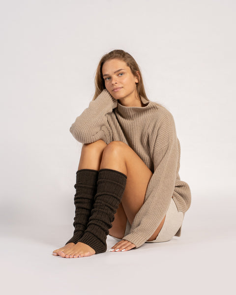 A woman sits on the floor in a studio setting, wearing an oversized beige mock-neck sweater and dark brown toeless leg warmers, looking at the camera.