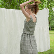 The model is wearing the matching set of the a-line SOPHIA skirt and the ANA crop top in green gingham.  She is brushing her hair with her hands, moving from side to side, while swinging her arms. She appears to be surrounded by trees in a garden.