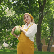 Young blond woman wearing Nicci linen jumpsuit in spicy yellow - eating watermelon and standing in the nature's landscape