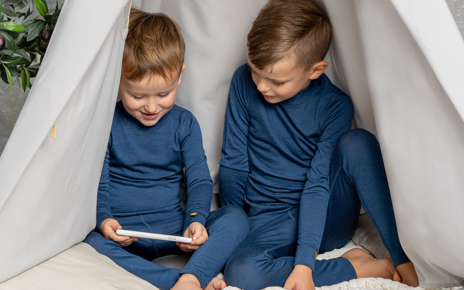 Two boys sitting inside a play tent, wearing the menique merino wool 160gsm base layers sets of long sleeve shirt and bottoms in denim color.