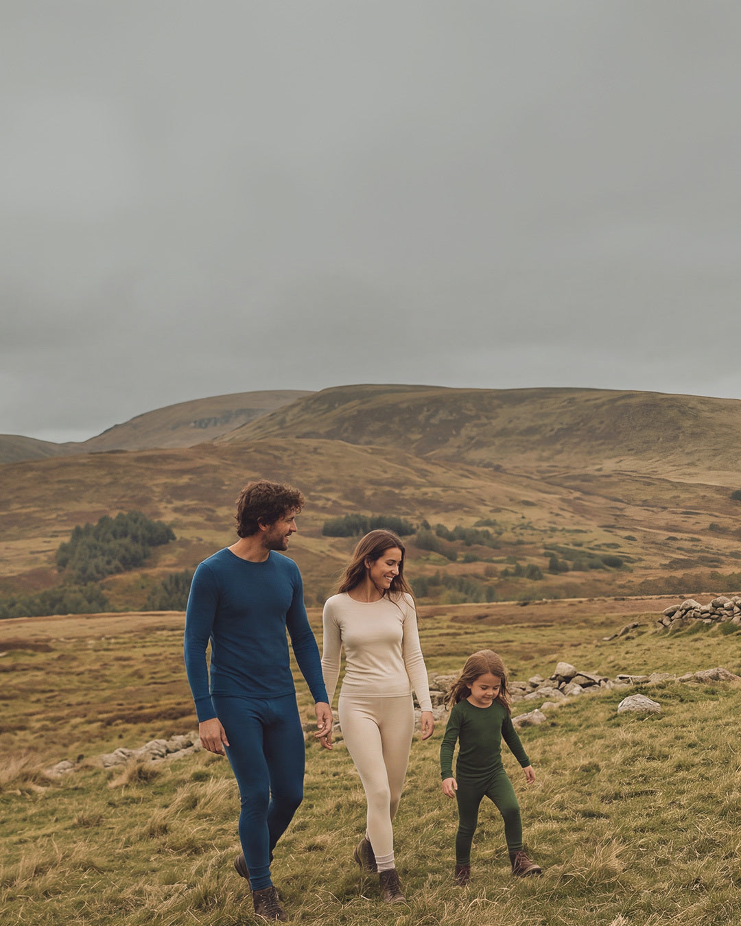 A man, woman, and young child walk toward the camera while holding hands in a rugged outdoor setting. The family is dressed in coordinating blue and beige outfits under an overcast, moody sky.