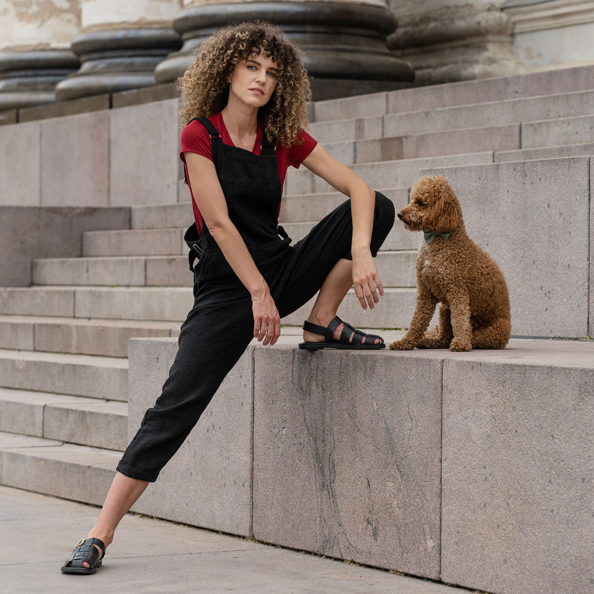 Beautiful woman with curly hair posing in the city wearing pure black linen jumpsuit Nicci, royal cherry merino wool top. Posing with a brown poodle dog.