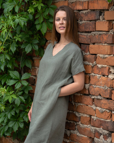 Young woman posing near the red brick wall, wearing stone green color linen tunic dress Joelle with her hand tucked in a pocket.