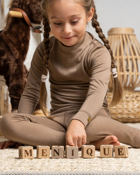 a girl wearing the menique Kids&#39; Merino 160 Long Sleeve Crew in Beige color. She is playing with wooden  letter cubes on the carpet.