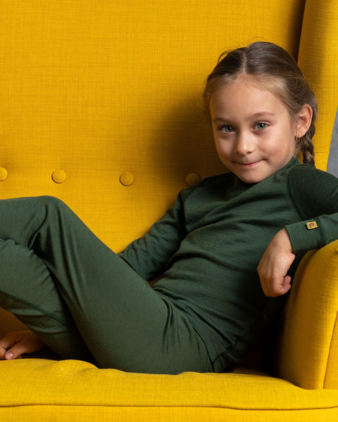 a girl, lying in a spicy yellow color ottoman, with her arm on the arm rest. She is wearing the menique Kids&#39; Long Sleeve Crew 160 Merino in Dark Green color.